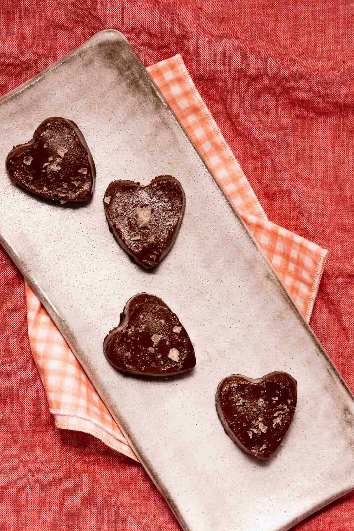 Four heart-shaped homemade chocolate rosemary pine nut truffles topped with flake salt on a small serving rectangle over pink linens.