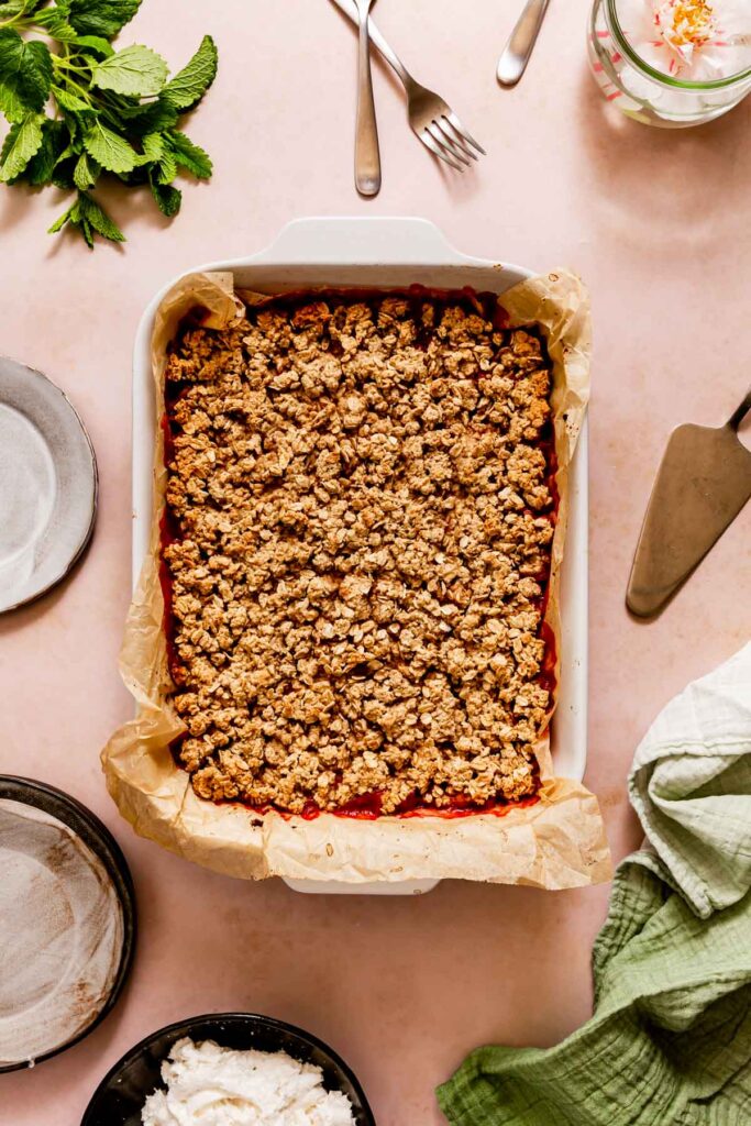 A 9x13 pan of strawberry rhubarb crisp surrounded by whipped cream, plates, and lemon balm on a pink background.