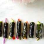 Tiny sushi rolls with colorful veggies inside lined up on a marble table with a pink peony next to them.