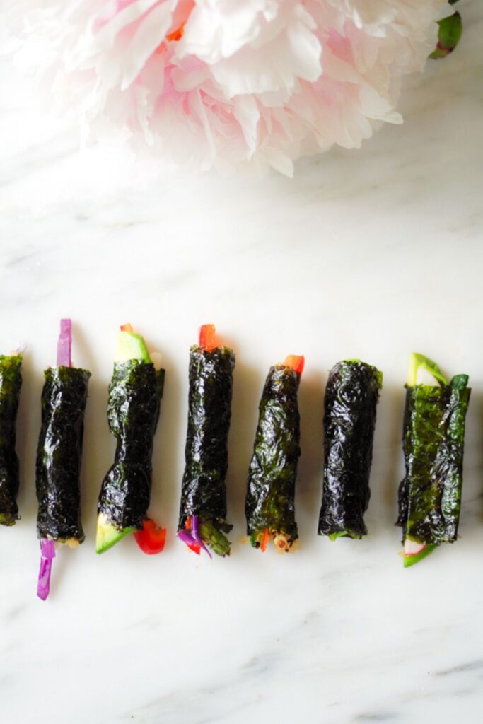 Tiny sushi rolls with colorful veggies inside lined up on a marble table with a pink peony next to them.