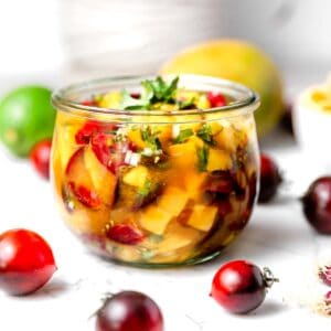 A rounded glass jar of freshly made mango salsa surrounded by cherry tomatoes, a lime, and a mango.