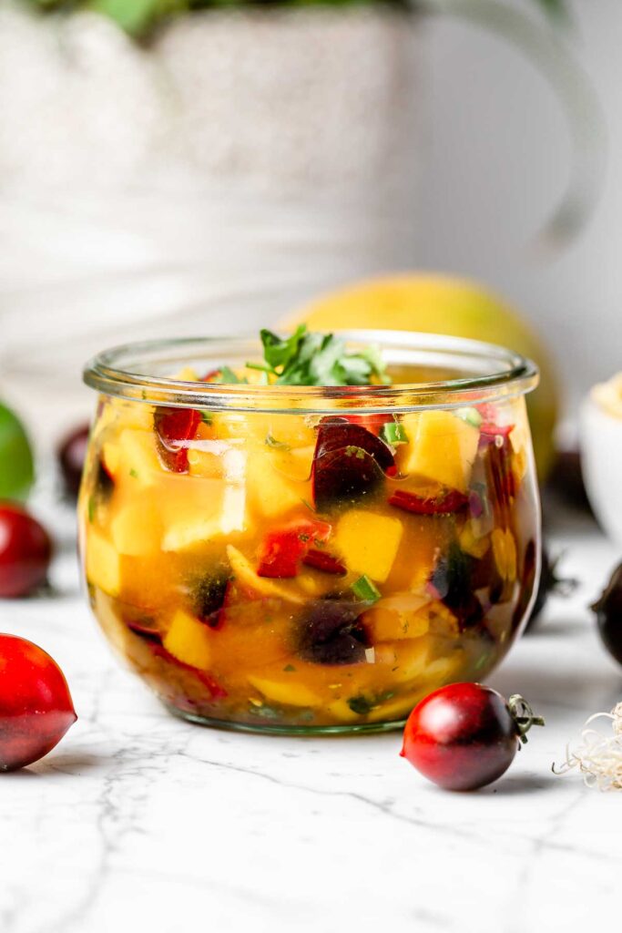 A round glass jar filled with mango salsa with cherry tomatoes surrounding it.