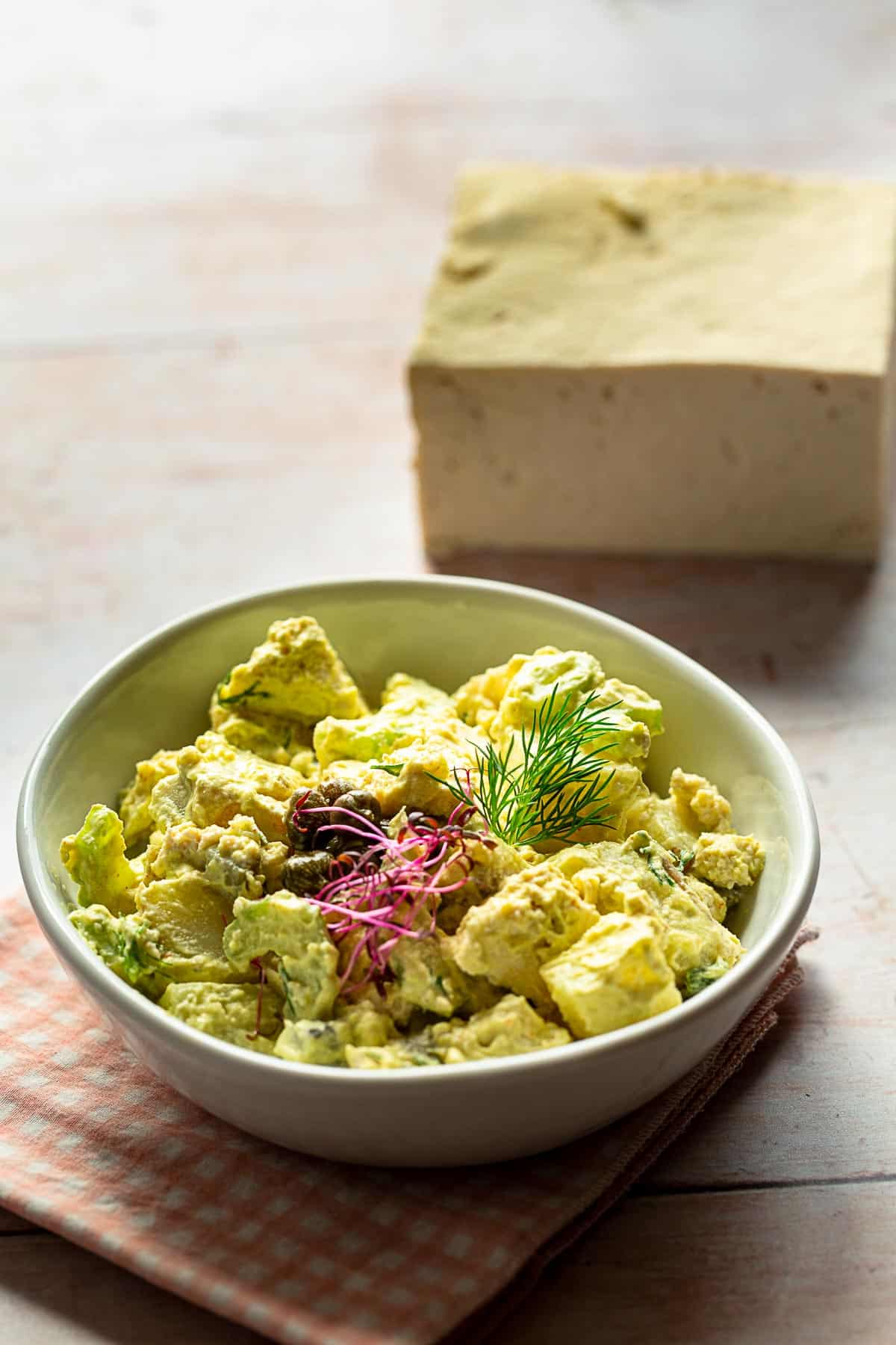 A small bowl of potato salad topped with pink micro greens with a block of tofu behind it.