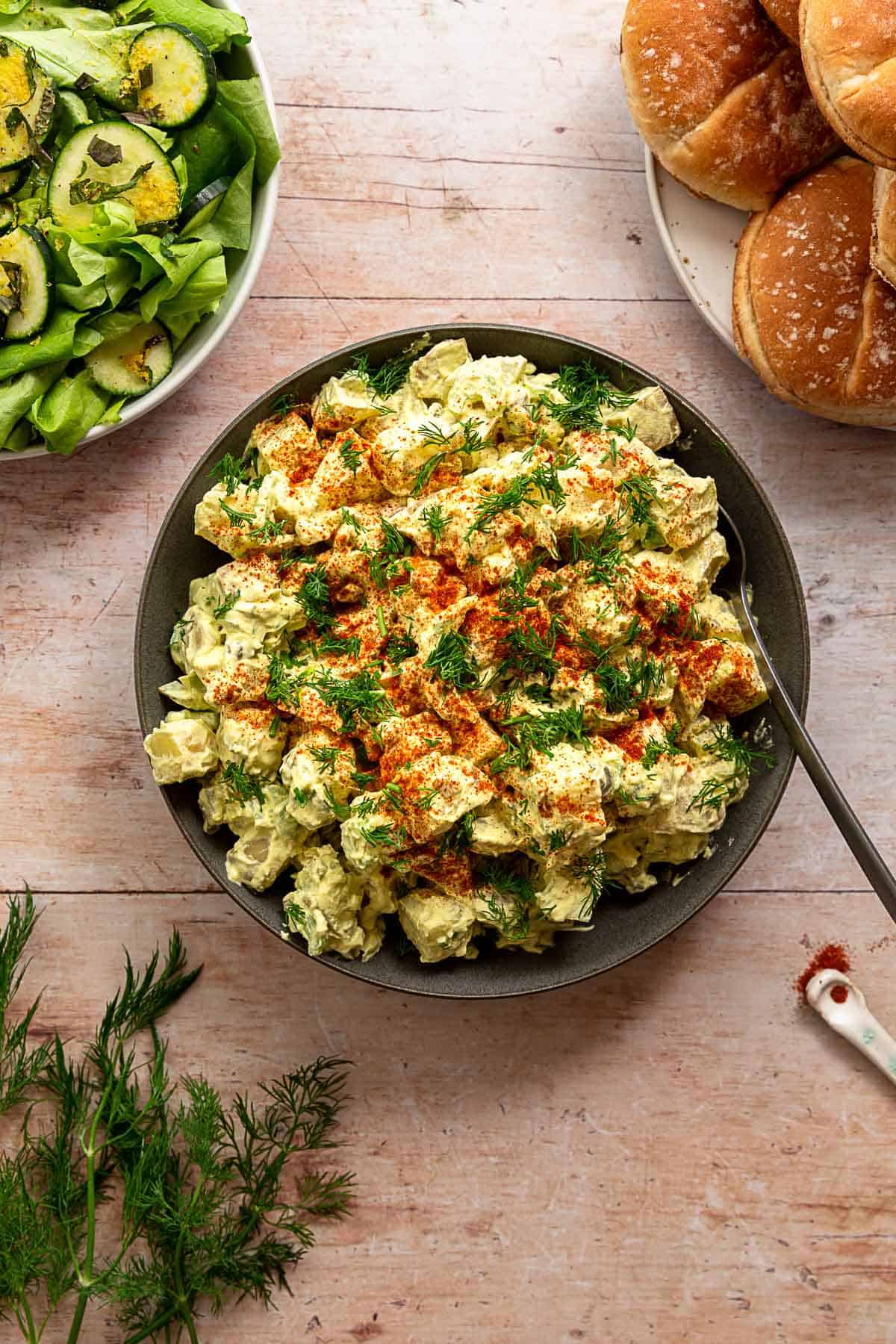 Classic potato salad with green salad, dill, and hamburger buns surrounding it on a wood surface.