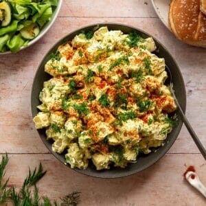 Classic potato salad veganized, topped with paprika and dill and surrounded by a green cucumber salad and hamburger buns.