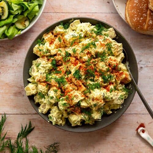 Classic potato salad veganized, topped with paprika and dill and surrounded by a green cucumber salad and hamburger buns.