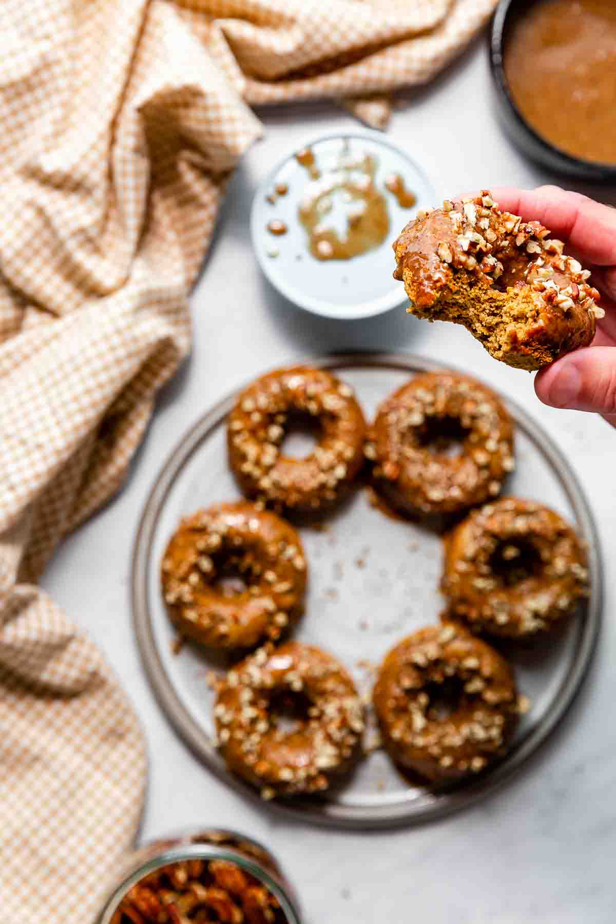 Baked vegan & gluten-free pumpkin donuts with pecan topping on a grey platter below a hand holding a bitten donut.