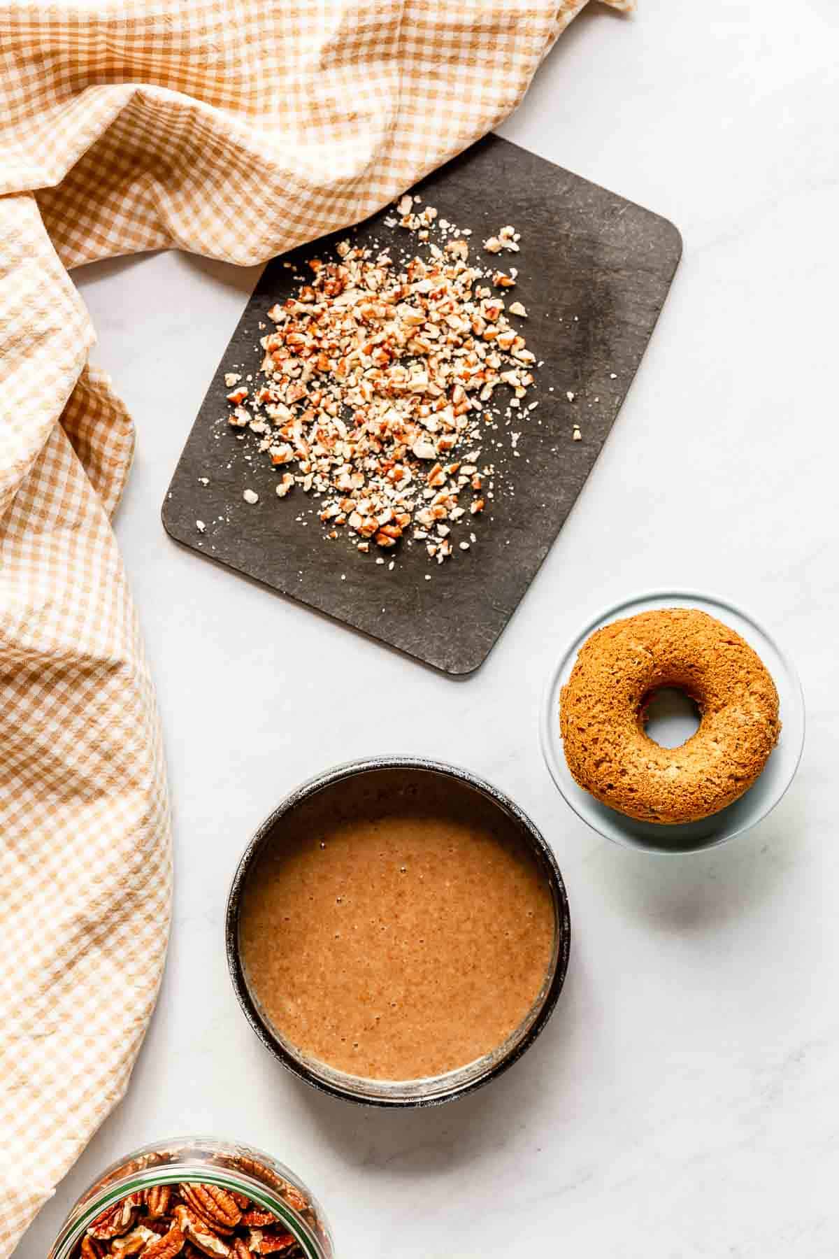 A freshly baked donut between a bowl of pecan caramel and a small black cutting board filled with chopped pecans.