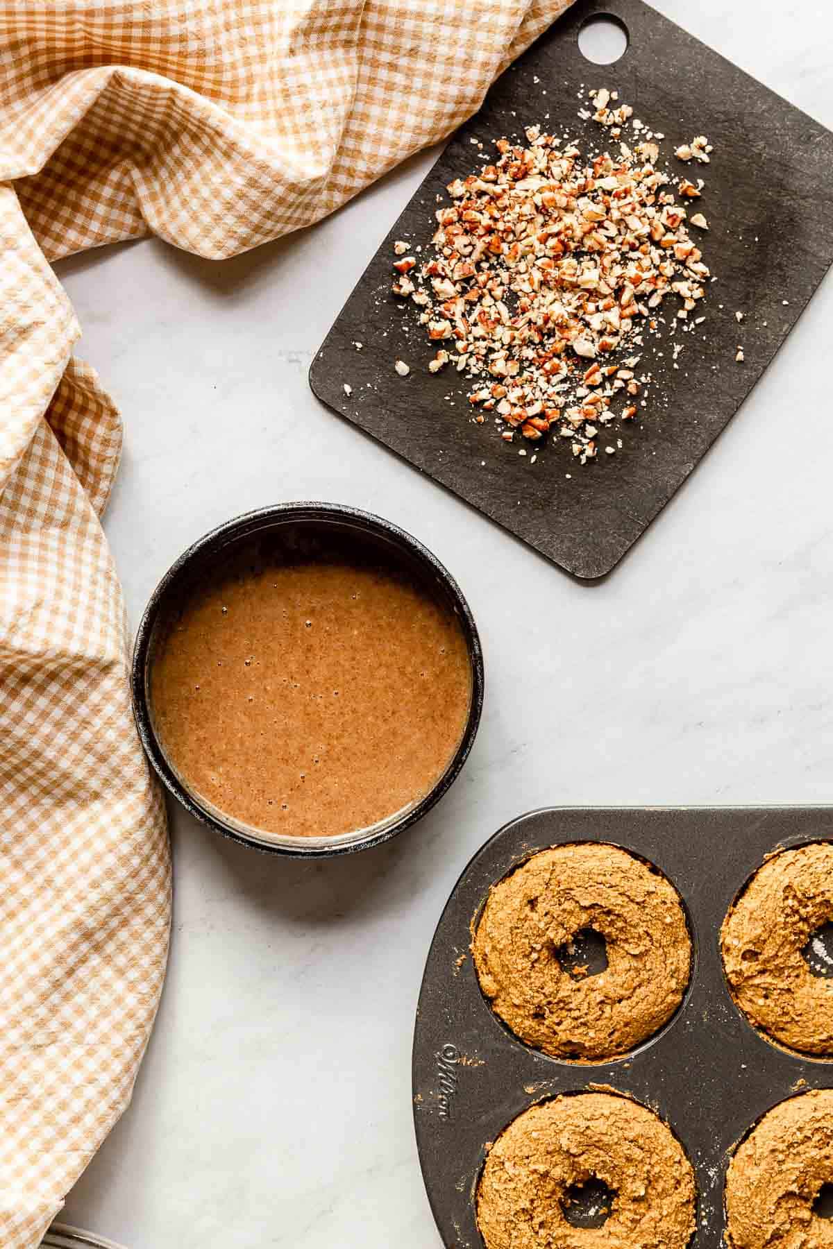 Fresh out of the oven baked pumpkin donuts in a donut pan next to a bowl of pecan caramel sauce and a small black cutting board filled with chopped pecans.