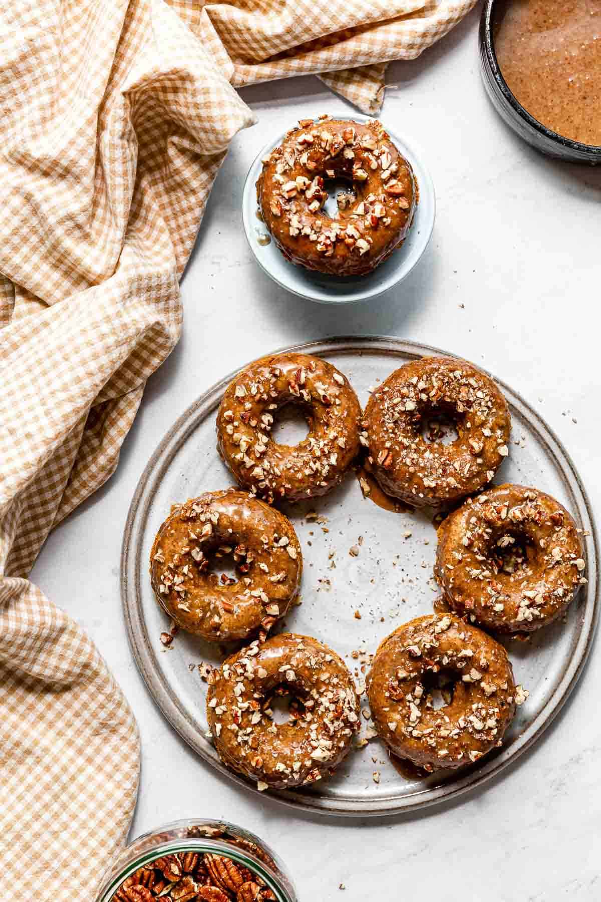 Vegan baked pumpkin doughnuts with pecan topping on a grey platter below a single doughnut on a small pedestal, surrounded by pecan caramel and whole pecans.