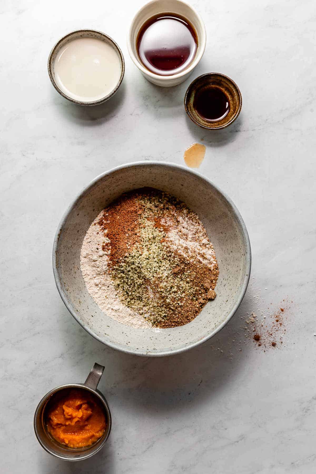 Dry ingredients for making homemade baked pumpkin donuts in a mixing bowl surrounded by the wet ingredients on a marble surface.