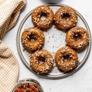 Vegan baked pumpkin donuts with pecan topping on a grey platter and a brown gingham cloth with a bowl of candied pecans below.