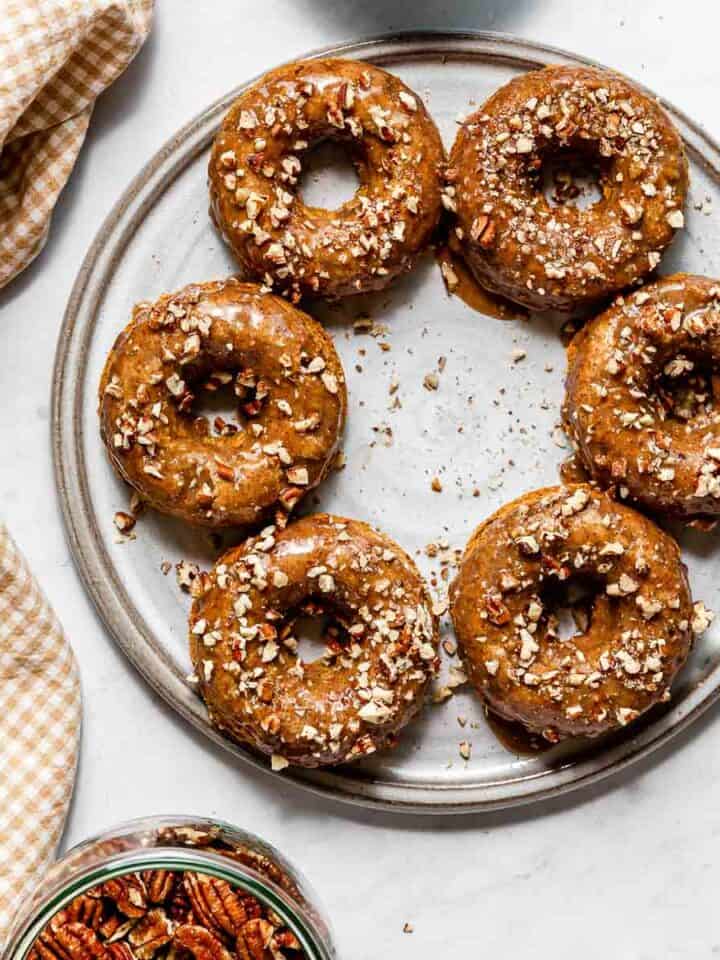 Vegan baked pumpkin donuts with pecan topping on a grey platter and a brown gingham cloth with a bowl of candied pecans below.