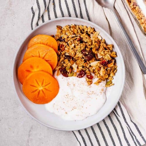 A breakfast bowl of oil-free orange pecan granola with persimmons and vegan yogurt.