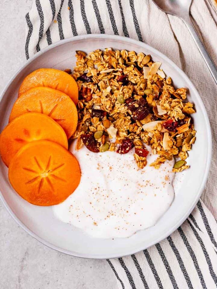 A breakfast bowl of oil-free orange pecan granola with persimmons and vegan yogurt.