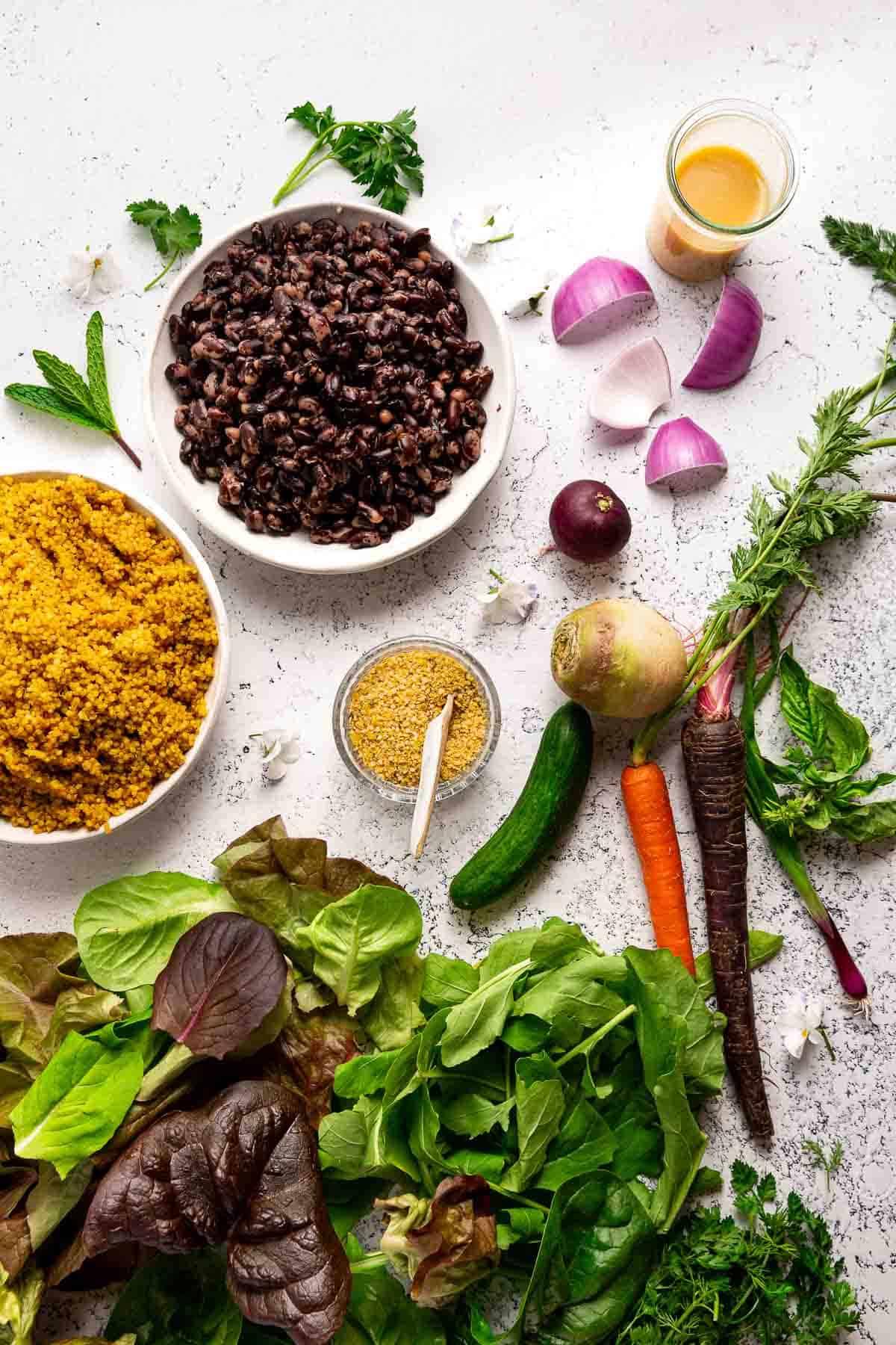 Greens, colorful veggies, turmeric-quinoa, black and white orca beans, herbs, seeds, and dressing for making vegan buddha bowls.