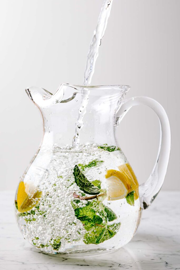 Mint sprigs and lemon slices floating in a glass pitcher with water pouring in.
