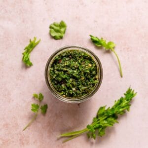 A jar of chimichurri surrounded by parsley and cilantro leaves.