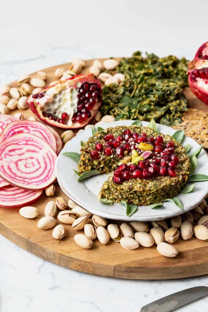 a vegan cheese board with nuts, candy cane beets, a green cheesy spread topped with pomegranates 
