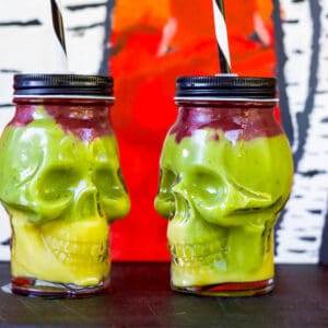Two vegan halloween smoothie treats colored yellow, green, and purple in skull-shaped jars.