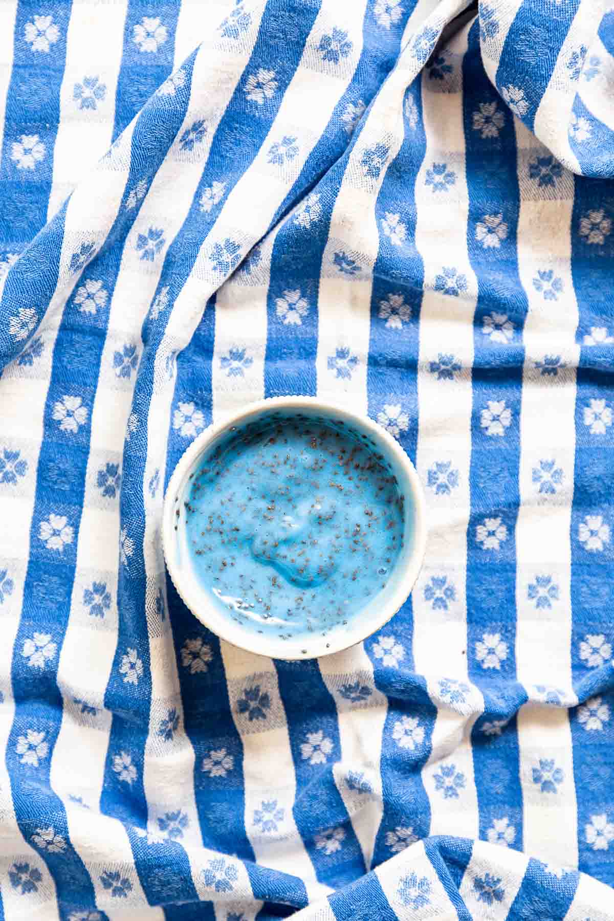 Blue yogurt with chia sees stirred into it, inside a ramekin on top of a blue gingham tablecloth.