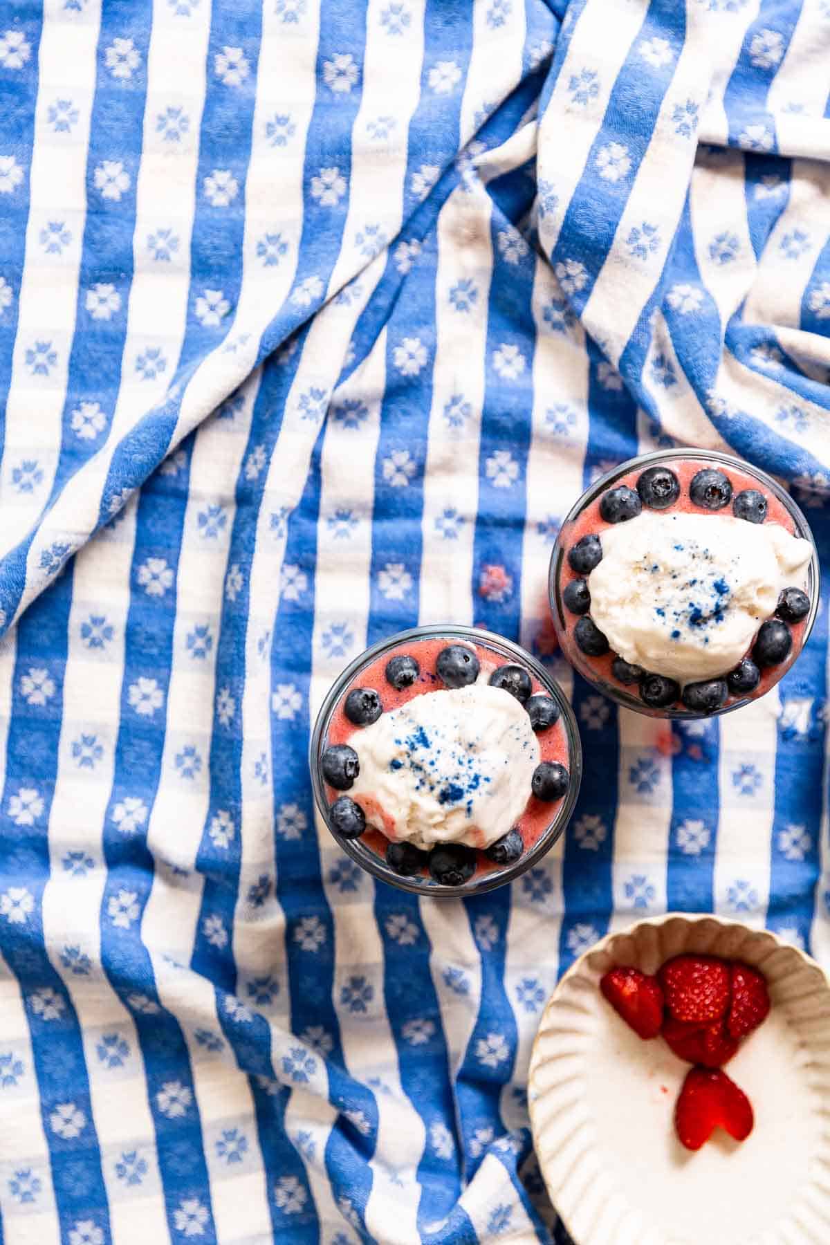 Blueberries, whipped cream, and blue powder on strawberry smoothie in jars atop a blue gingham tablecloth.