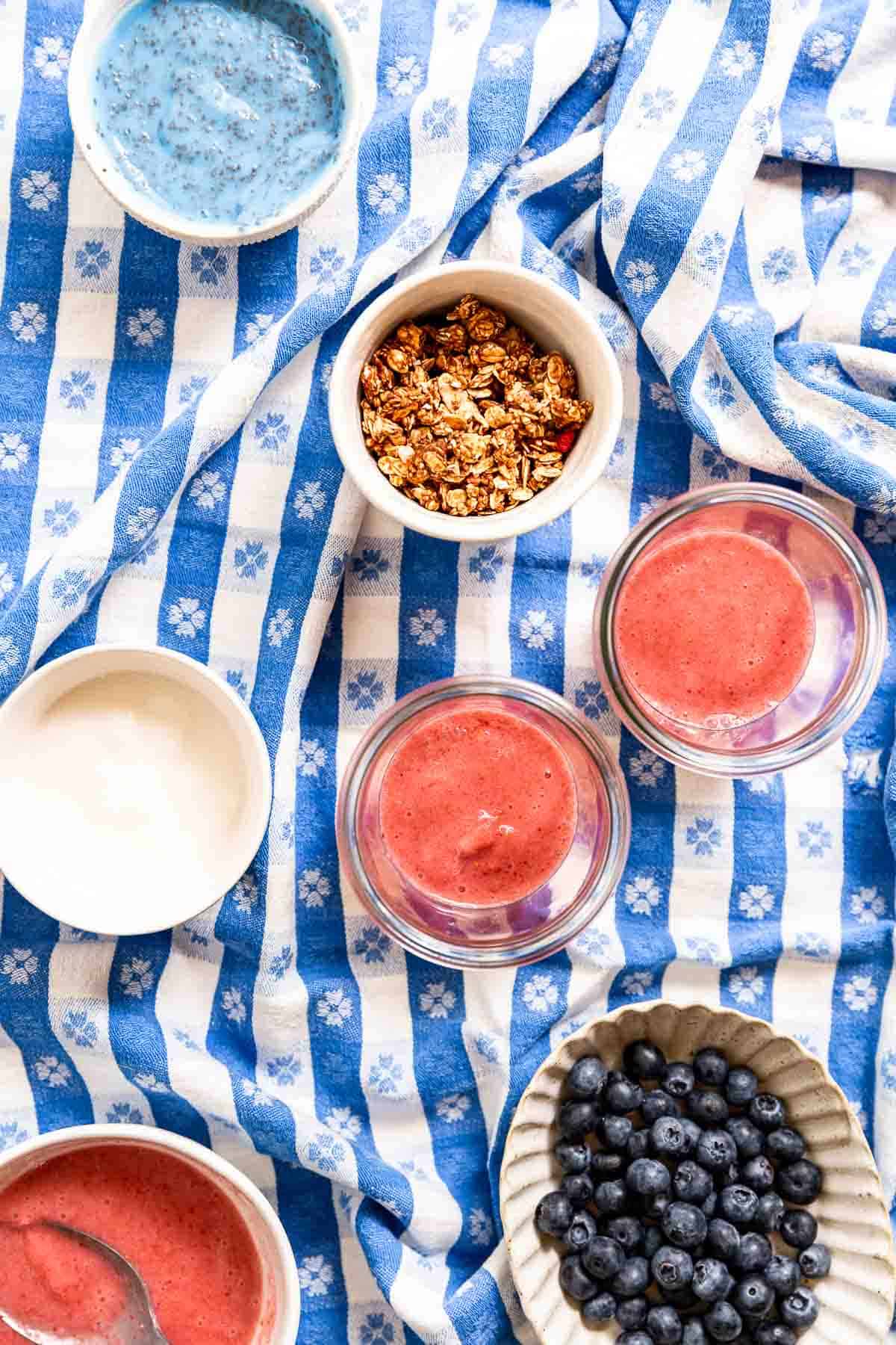 Two jars with strawberry smoothie inside surrounded by ramekins of granola, yogurt, and blueberries.