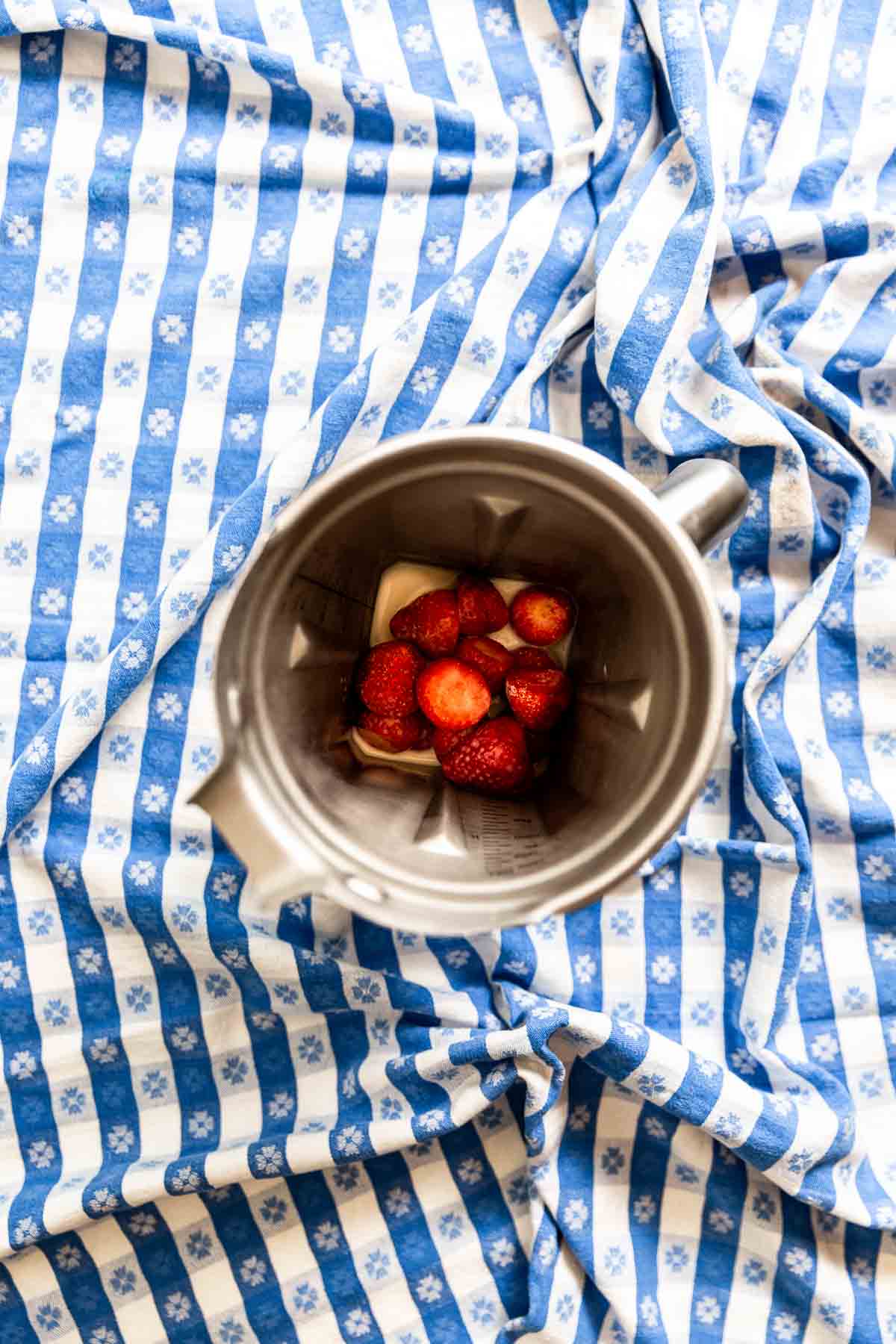 A stainless steel blender filled with frozen strawberries on a blue gingham tablecloth.