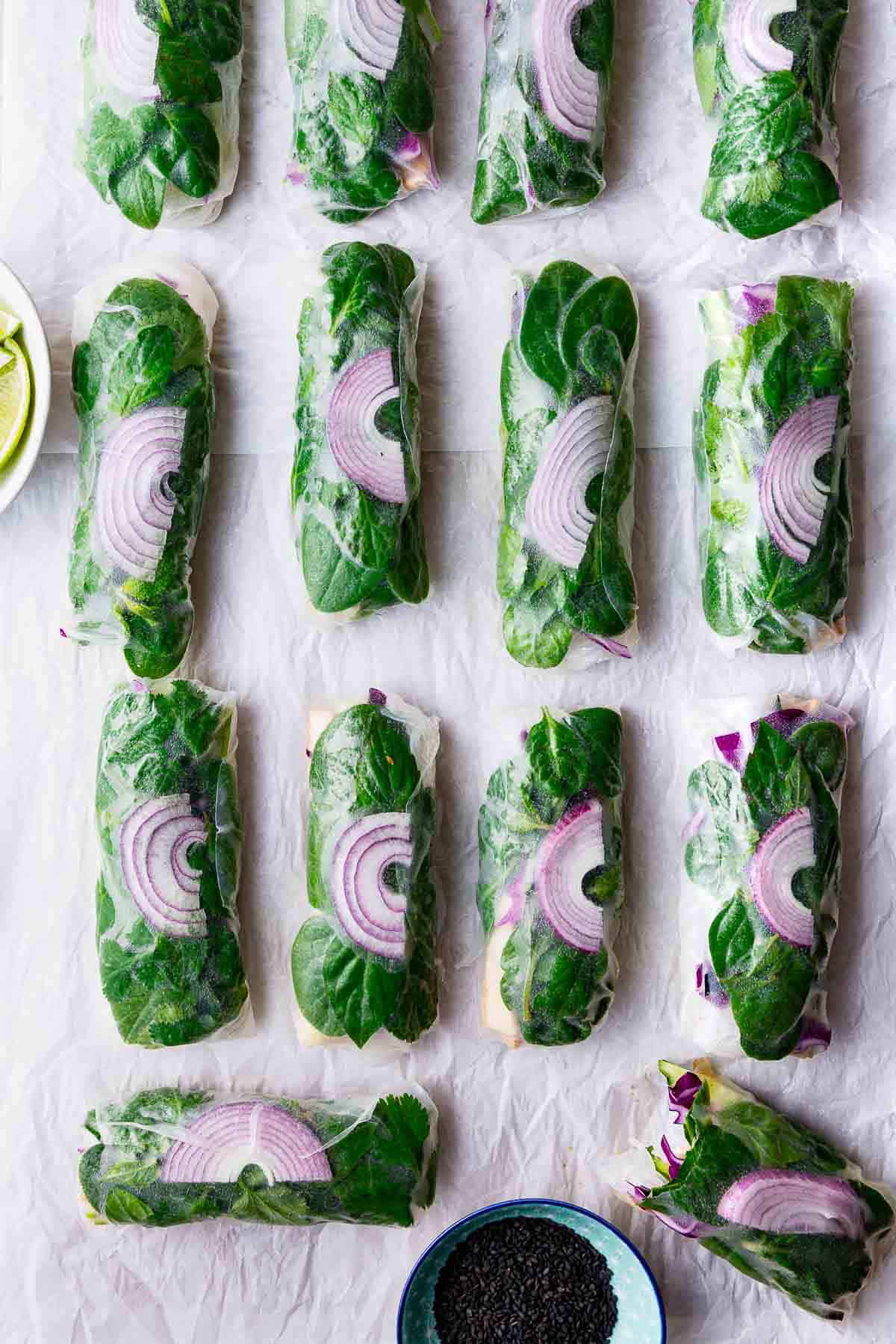 An overhead view of 14 green and purple summer rolls on white parchment, with bowls of dipping sauce and limes next to them.