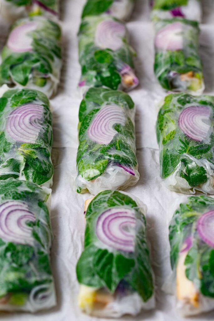 On white parchment, rows of summer rolls with greens and purple onion slices inside.