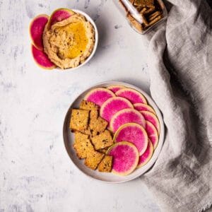 A platter of homemade artisanal rosemary olive crackers with sliced watermelon radishes and hummus.