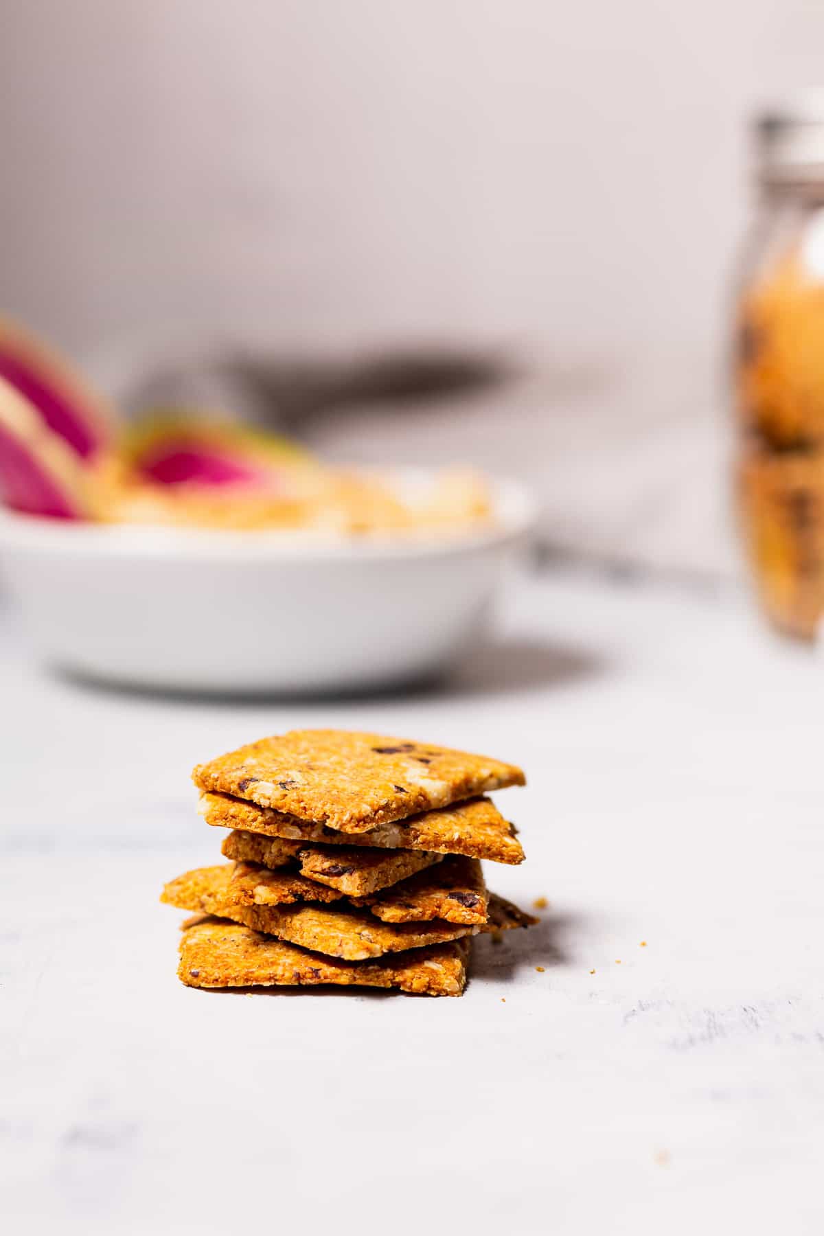 A stack of homemade rosemary olive crackers in front of a bowl of hummus and a jarful of crackers.