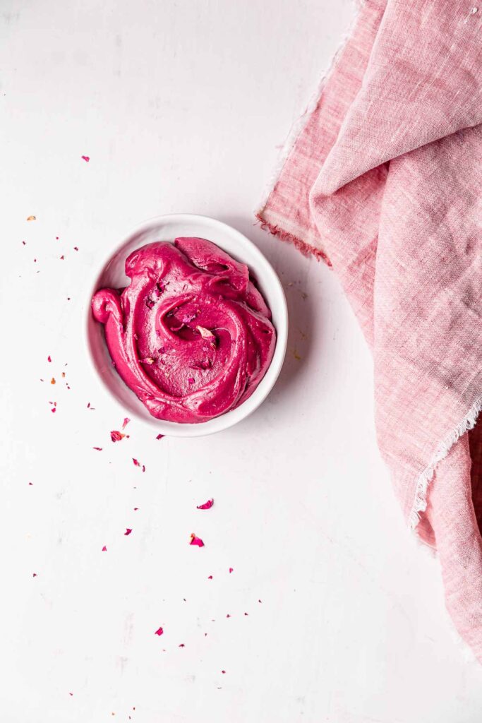 A bowl of vibrant pink frosting surrounded by rose petals and a pink linen.