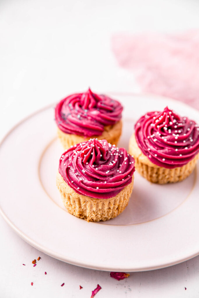 Three vanilla cupcakes topped with vibrant pink frosting on a plate. 