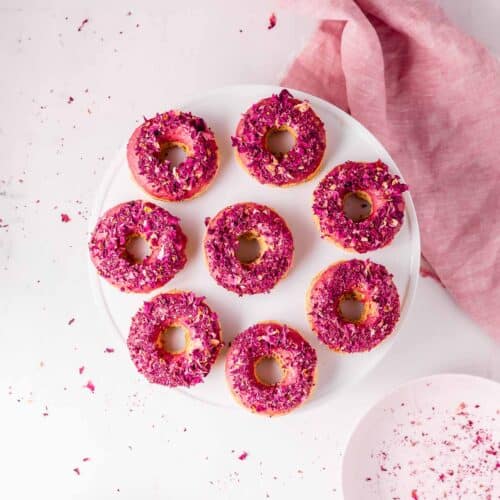 A topdown view of a white cake stand filled with a ring of pink donuts topped with rose petals.