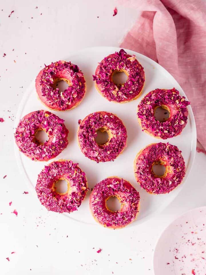 A topdown view of a white cake stand filled with a ring of pink donuts topped with rose petals.