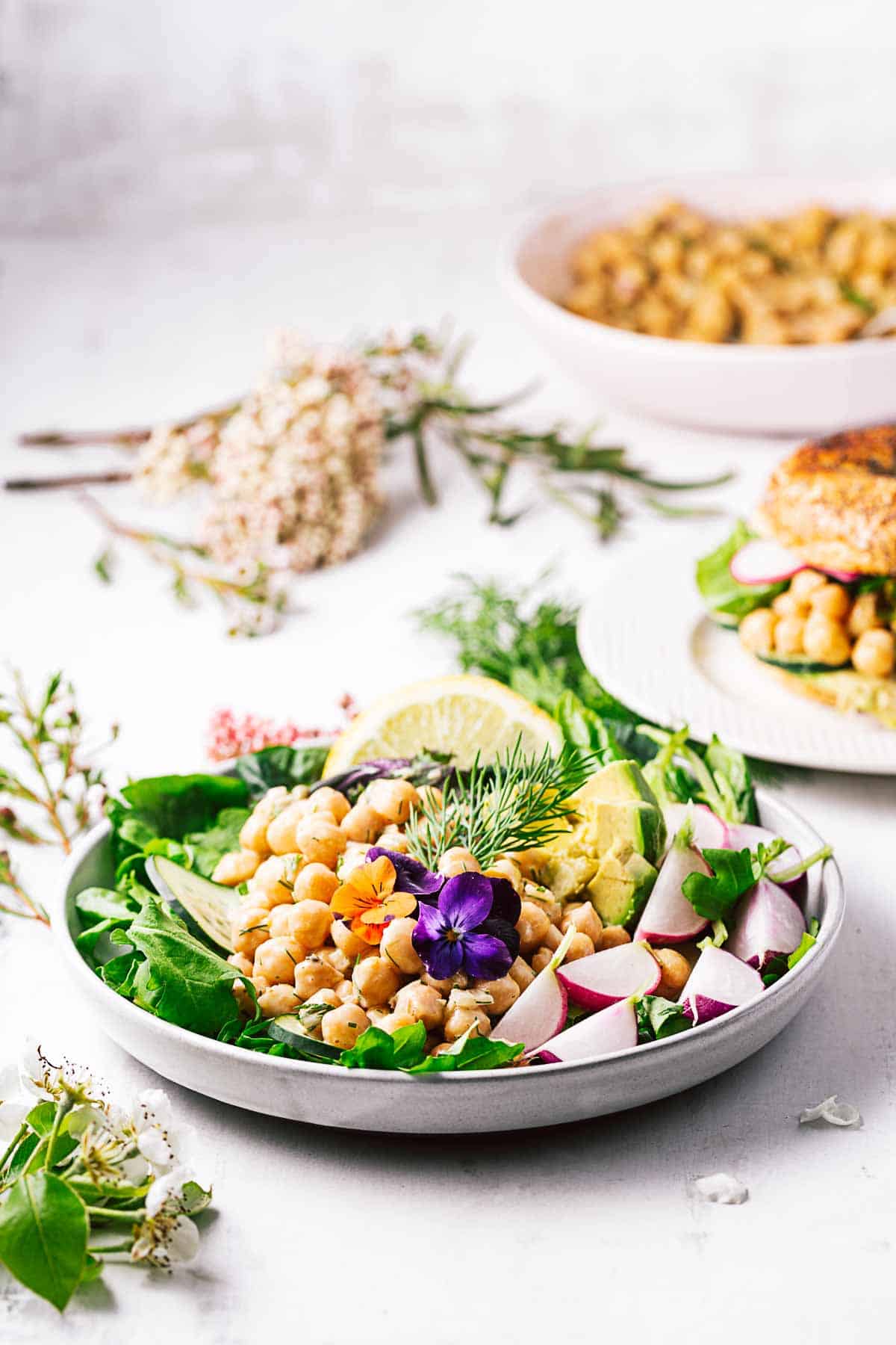 A salad bowl in the foreground with chickpea salad on top, a bagel sandwich filled with vegan chickpea salad behind, and a bowl of just vegan chickpea salad behind that. 