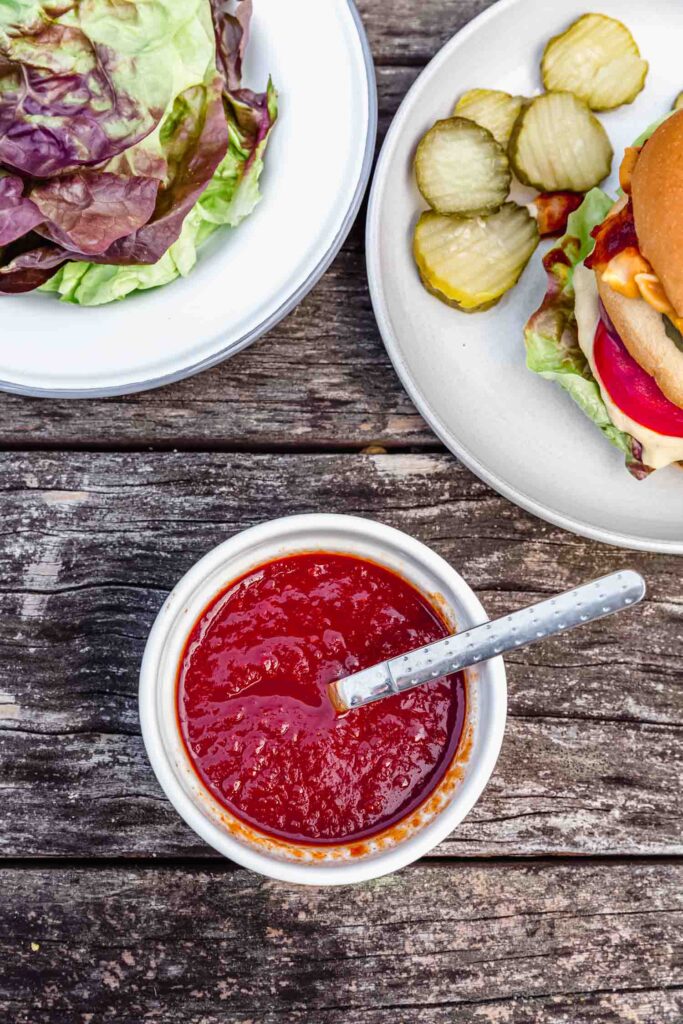 A ramekin filled with bbq sauce surrounded by plates of lettuce and burgers.