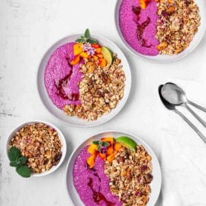 Three tropical breakfast bowls with pink dragon fruit chia pudding, red jam, tropical granola, and mango pieces.