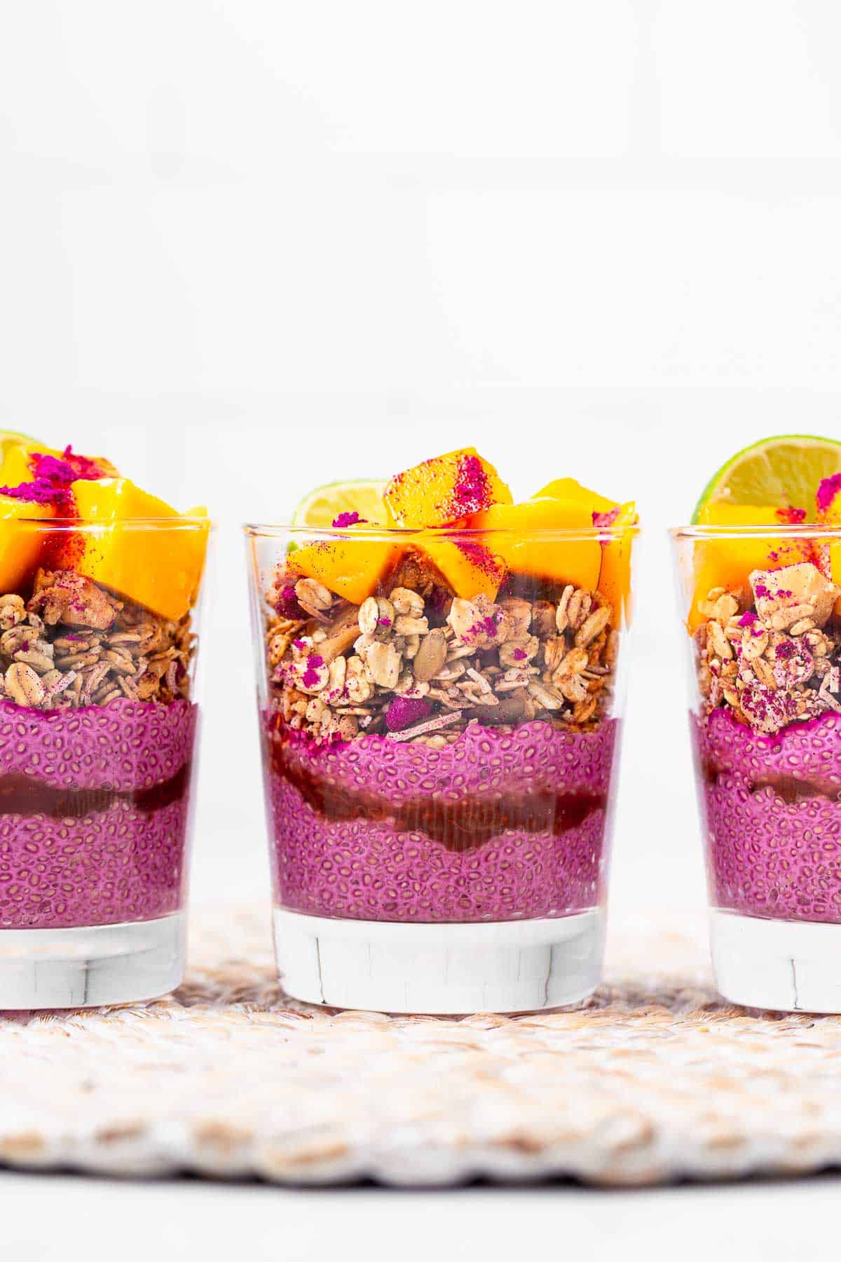 A side view of three glasses layered with pink chia pudding, red jam, tropical granola, and mango chunks.