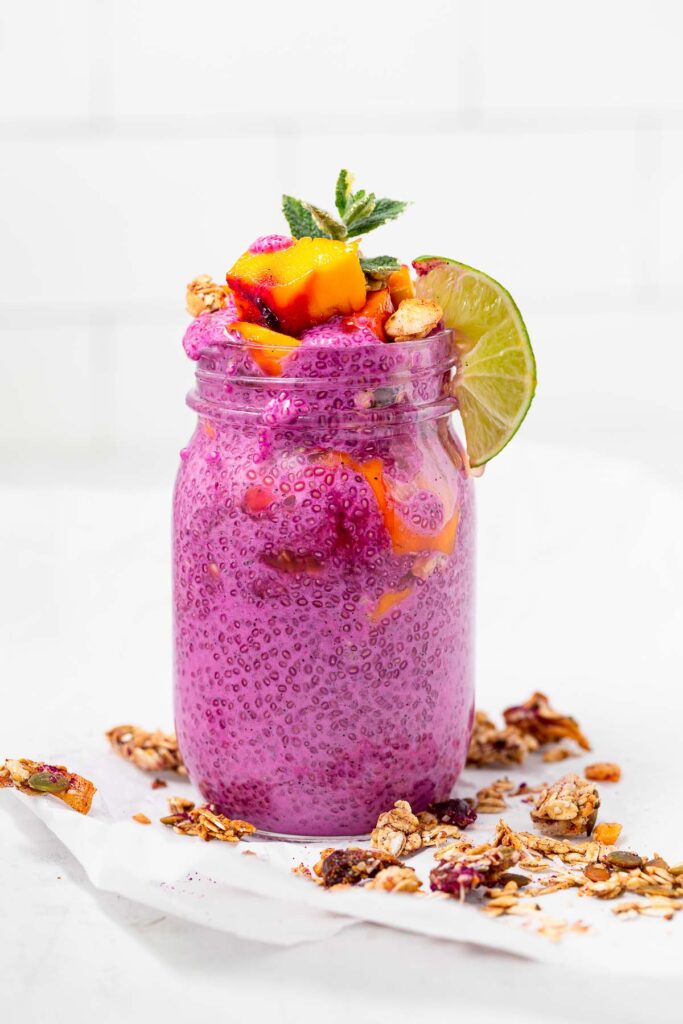 A large mason jar filled with pink dragon fruit chia pudding, topped with mango and mint and surrounded by tropical granola.