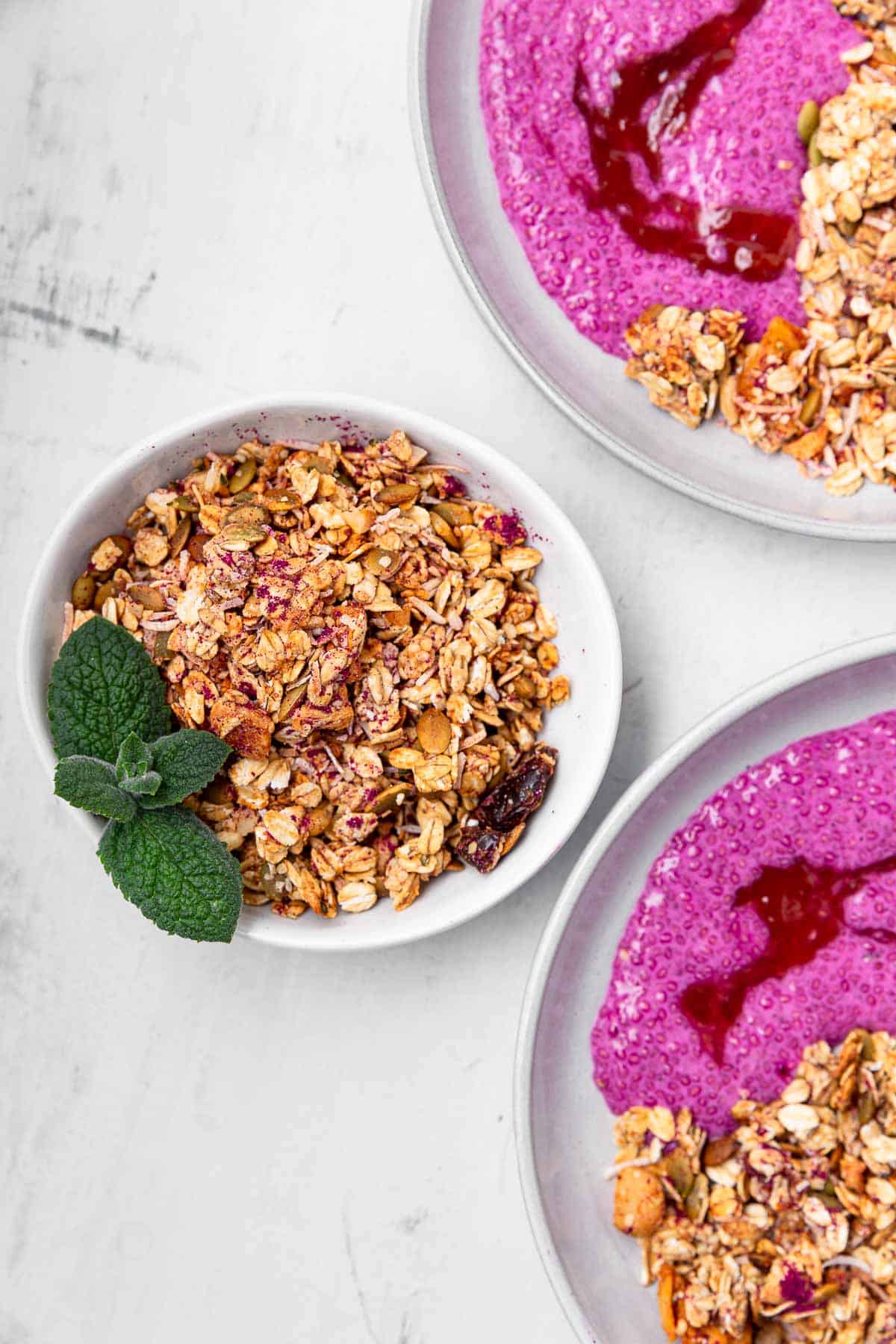 A bowl of tropical granola garnished with mint next to two breakfast bowls filled with the granola and pink chia pudding.