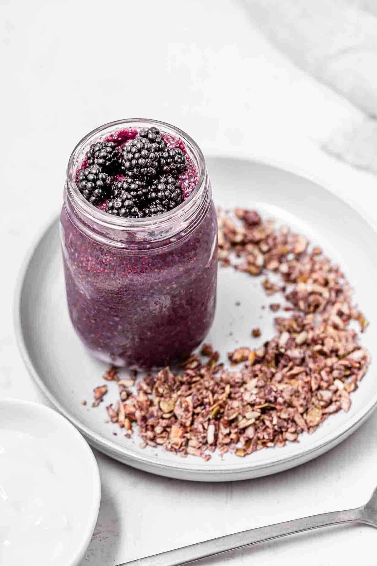 A jar of blackberry chia pudding on a grey plate filled with granola.