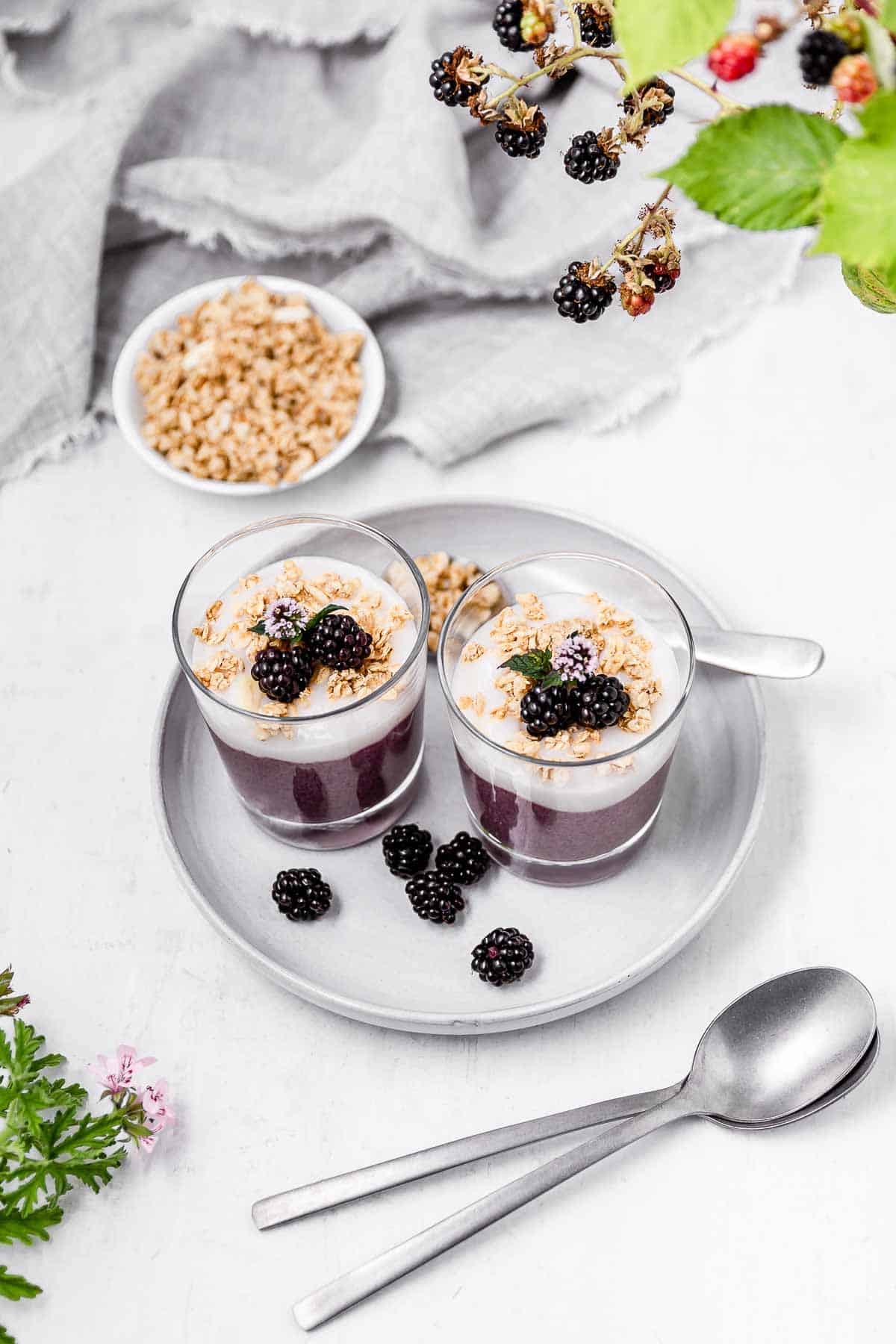 Two small glass serving jars filled with blackberry chia pudding topped with yogurt and granola, all on a grey plate with fresh blackberries on it and two spoons next to it.