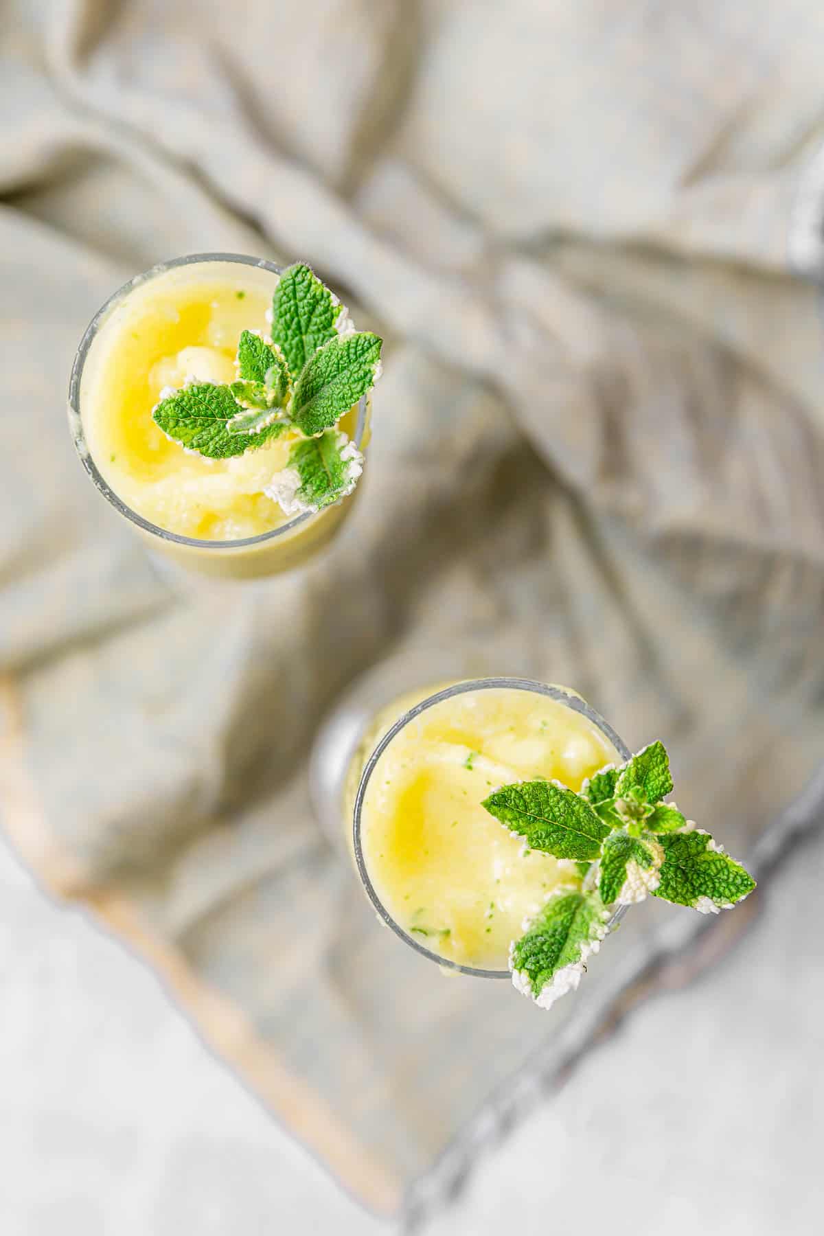An overhead view of two pineapple mint frozen drinks in tall glasses, topped with mint.