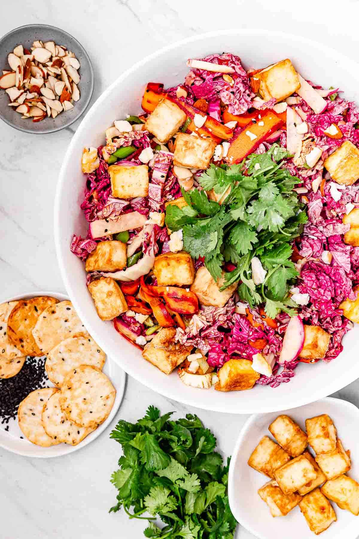 An overhead view of a big salad bowl filled with colorful sesame tofu salad, surrounded by slivered almonds, sesame crackers, cilantro, and crispy tofu.