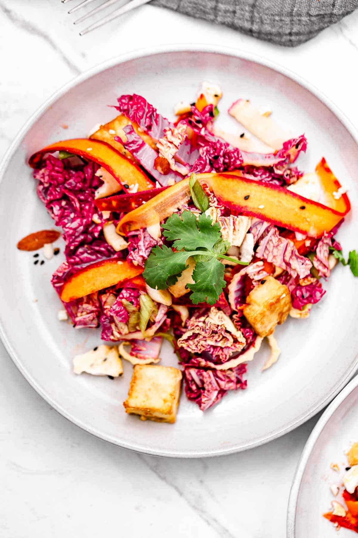 A closeup overhead view of a salad made of red napa cabbage, colorful carrots, sesame, and tofu.