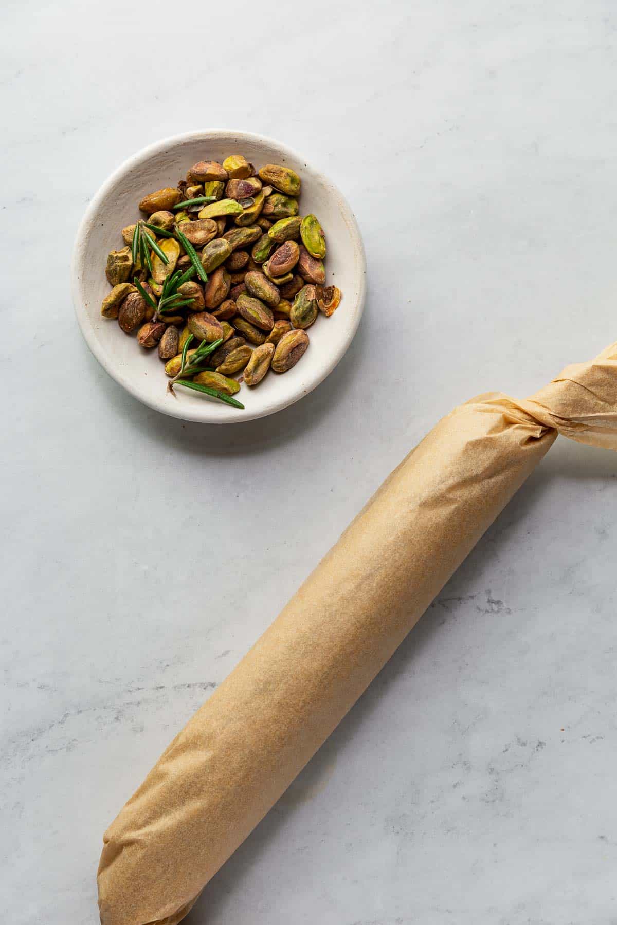 A Lemon Rosemary Vegan Cheese Log rolled up in parchment next to a bowl of pistachios for the topping.