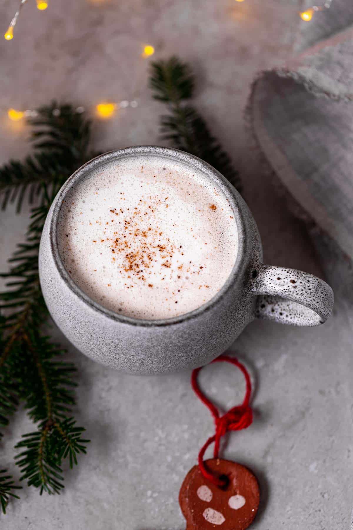 A large mug of dairy-free eggnog topped with whipped cream and nutmeg, surrounded by lights, branches, and a gingerbread ornament.