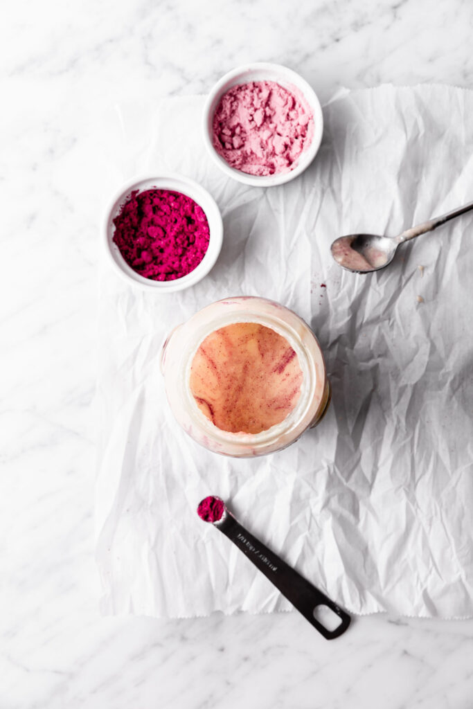 An overhead view of a jar of white nut butter with pink fruit powders in bowls next to it.